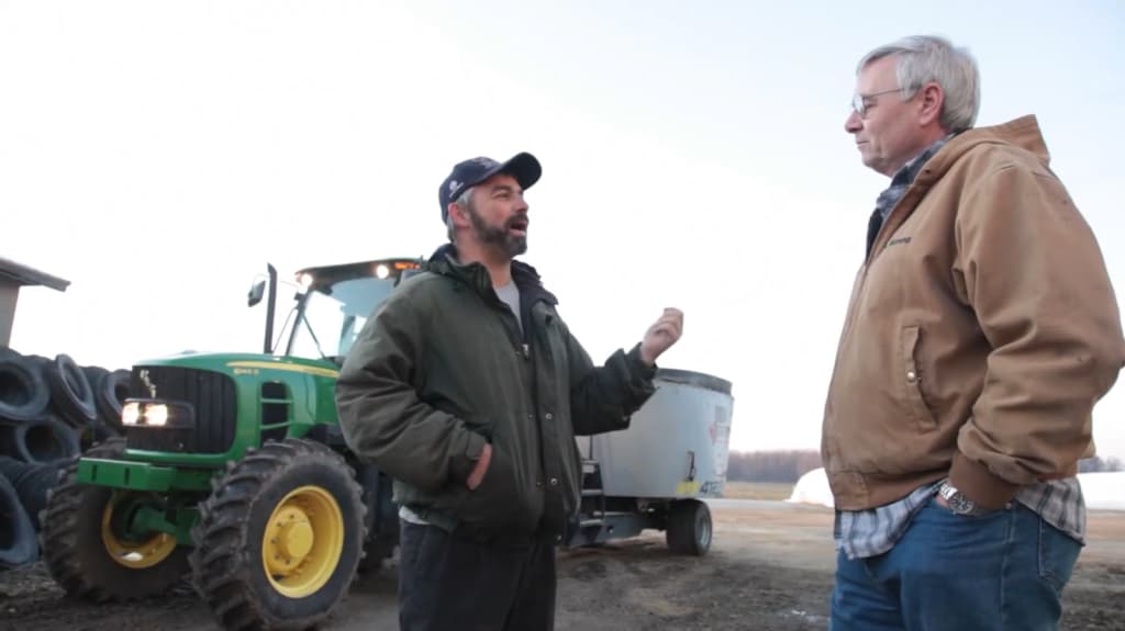 Outdoors, the man talking to another man beside a green tractor