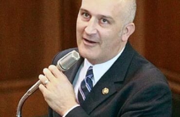 A man in a dark suit and tie speaking into a handheld microphone