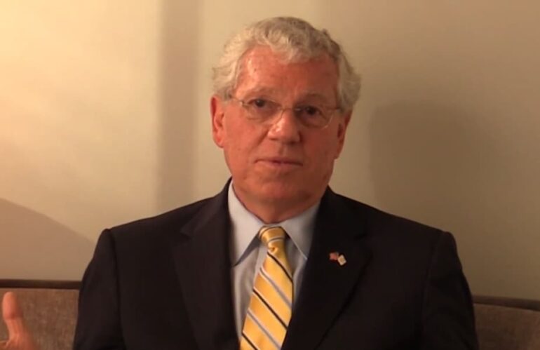 A man with grey hair, glasses, and a striped tie speaks in an interview