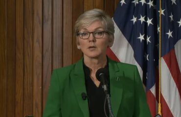 A woman in a green blazer speaks at a podium with a US flag behind her