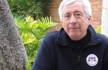 Joe Adame smiling seated outdoors, wearing a jacket with a badge