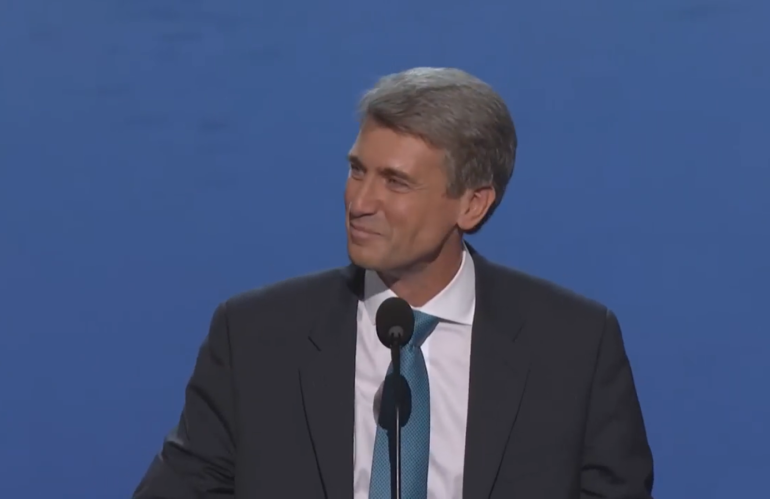 A smiling man in a suit stands at a podium in front of a blue wall