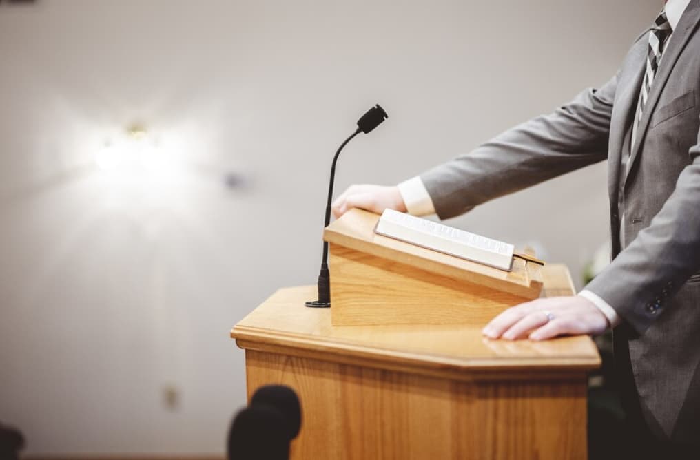 A person stands at a wooden podium with a microphone and a book