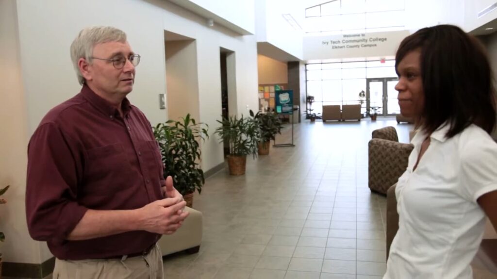 A man in glasses speaks to a woman in a college lobby